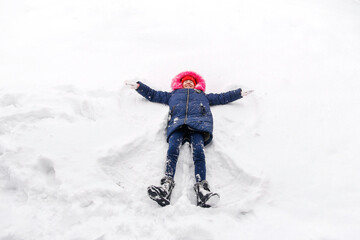 Fototapeta na wymiar Preteen smiling girl in bright pink warm hood making snow angel while lying on snow outside background. Pretty child outdoor, cold weather. Caucasian kid. Fun holidays and vacations. Winter enjoy