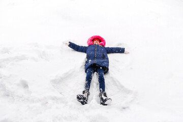 Fototapeta na wymiar Preteen smiling girl in bright pink warm hood making snow angel while lying on snow outside background. Playful child outdoor, cold weather. Caucasian kid. Fun holidays and vacations. Winter enjoy