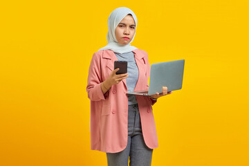 Angry and unhappy young Asian woman holding mobile phone and laptop on blue background