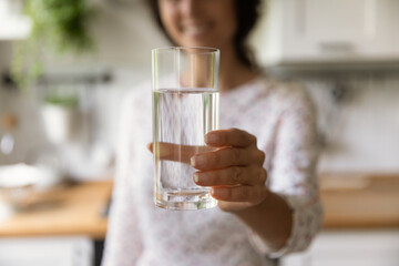 Happy woman holding glass of fresh filtered natural water in kitchen, toasting at camera, keeping...