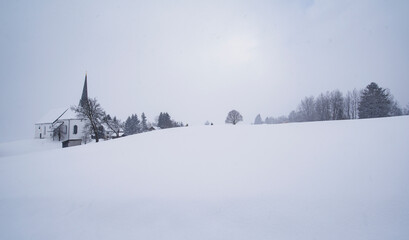 Kalte und verschneite Winterlandschaft und kleines Dorf mit Kirche in den bayerischen Alpen an einem stürmischen Tag 