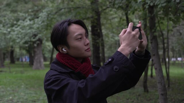 Young Asian man takes photo or video using smartphone in autumn park. The man takes pictures of nature on the phone