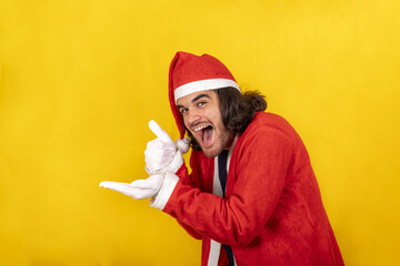 A smiling young man dressed as Santa Claus points at your object with his hand and raises his thumb in approval. Yellow background and copy space for your message. - obrazy, fototapety, plakaty