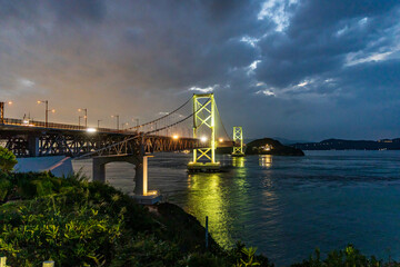 Naklejka premium Lights from suspension bridge mix with sunset glow and dramatic clouds