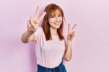 Redhead young woman wearing casual pink t shirt smiling looking to the camera showing fingers doing victory sign. number two.