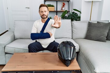 Middle age man with beard wearing arm on sling because motorbike accident with open hand doing stop...