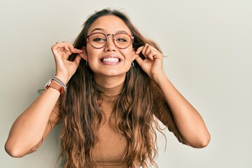 Young hispanic girl wearing casual clothes and glasses smiling pulling ears with fingers, funny...