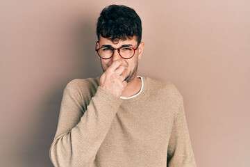 Fototapeta premium Young hispanic man wearing casual clothes and glasses smelling something stinky and disgusting, intolerable smell, holding breath with fingers on nose. bad smell
