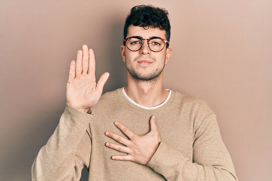 Young hispanic man wearing casual clothes and glasses swearing with hand on chest and open palm, making a loyalty promise oath