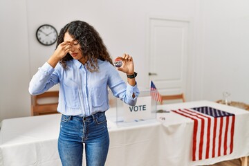 Beautiful hispanic woman standing by at political campaign by voting ballot tired rubbing nose and...
