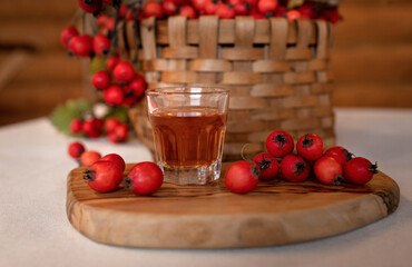A small shot glass with hawthorn tincture on the background of a wicker brown basket and scattered berries of ripe red hawthorn, a medicinal drink.