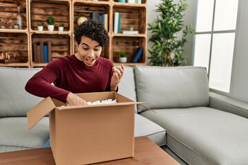 Young hispanic man unboxing cardboard box sitting on the sofa at home.