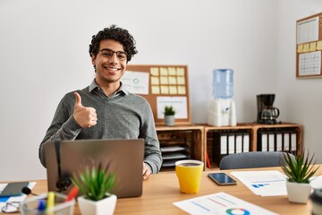 Fototapeta na wymiar Young hispanic man wearing business style sitting on desk at office doing happy thumbs up gesture with hand. approving expression looking at the camera showing success.