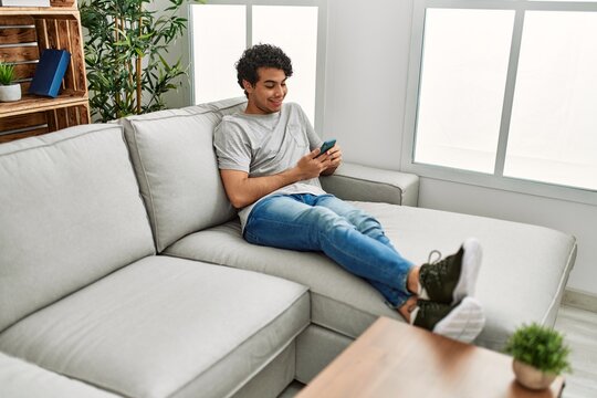 Young Hispanic Man Using Smartphone Sitting On The Sofa At Home.