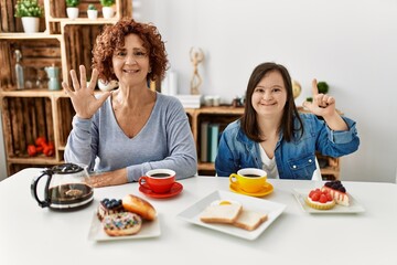 Family of mother and down syndrome daughter sitting at home eating breakfast showing and pointing up with fingers number seven while smiling confident and happy.