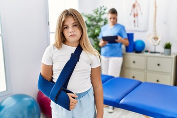 Blonde little girl wearing arm on sling at rehabilitation clinic thinking attitude and sober expression looking self confident