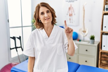 Fototapeta premium Middle age physiotherapist woman working at pain recovery clinic showing and pointing up with finger number one while smiling confident and happy.
