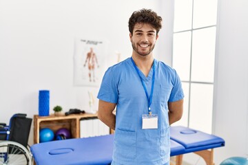 Young hispanic man wearing physio therapist uniform standing at clinic