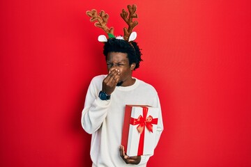 Young african american man wearing deer christmas hat holding gift smelling something stinky and...