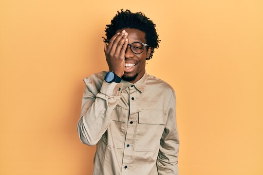Young african american man wearing casual clothes and glasses covering one eye with hand, confident smile on face and surprise emotion.