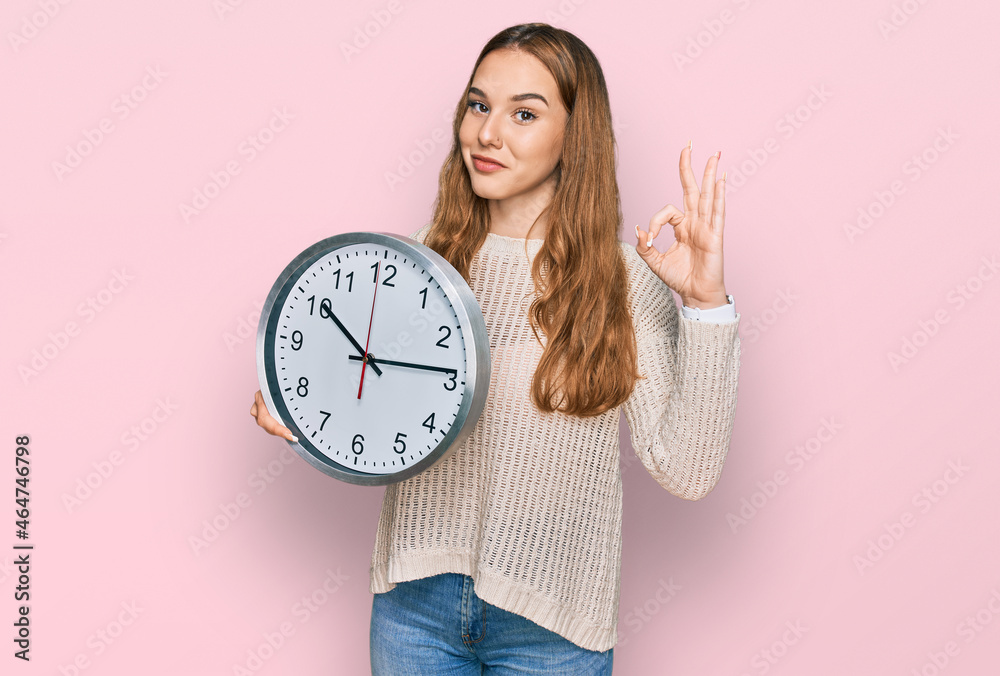 Sticker Young blonde woman holding big clock doing ok sign with fingers, smiling friendly gesturing excellent symbol