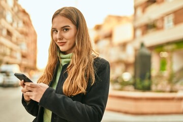 Young blonde girl smiling happy using smartphone at the city.