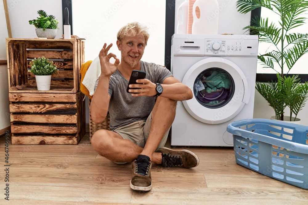 Poster Young blond man doing laundry using smartphone smiling positive doing ok sign with hand and fingers. successful expression.