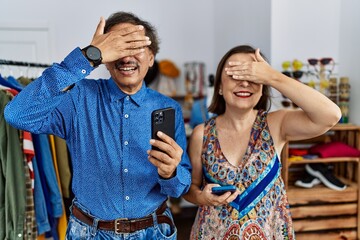 Middle age interracial couple at retail shop using smartphone smiling and laughing with hand on face covering eyes for surprise. blind concept.