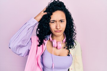 Young hispanic woman with curly hair wearing gym clothes and using headphones confuse and wonder about question. uncertain with doubt, thinking with hand on head. pensive concept.