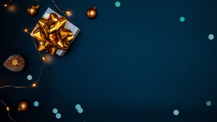 Christmas gift. White gift with golden bow, gold balls and sparkling lights garland in xmas decoration on dark background for greeting card. Xmas backdrop with space for text.