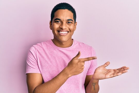 Young handsome hispanic man wearing casual pink t shirt amazed and smiling to the camera while presenting with hand and pointing with finger.