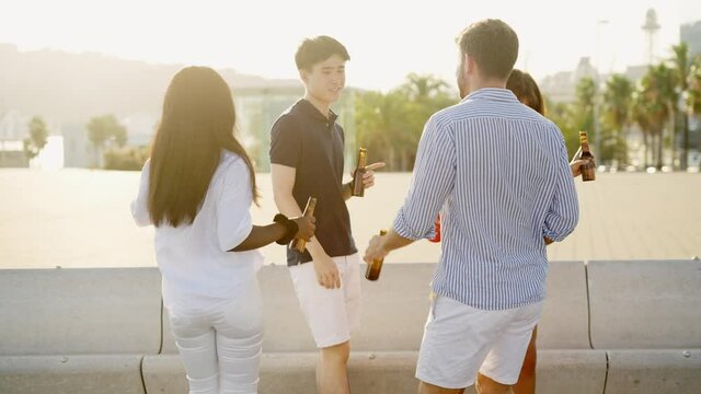 Group of young positive diverse friends enjoying summer sunset outdoors, dancing and drinking beer