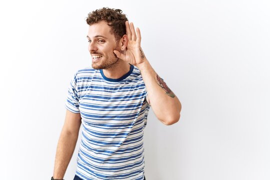 Handsome young man standing over isolated background smiling with hand over ear listening an hearing to rumor or gossip. deafness concept.