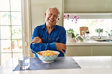 Senior man with grey hair eating pasta spaghetti at home happy face smiling with crossed arms looking at the camera. positive person.