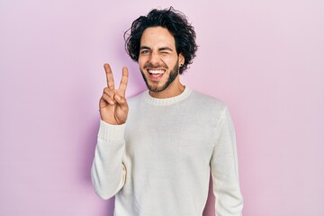 Fototapeta premium Handsome hispanic man wearing casual white sweater smiling with happy face winking at the camera doing victory sign with fingers. number two.