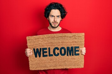 Handsome hispanic man holding welcome doormat skeptic and nervous, frowning upset because of...