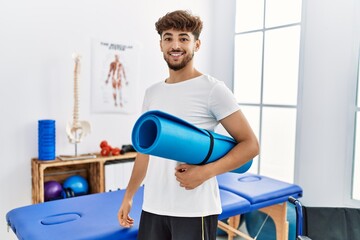 Young arab man patient smiling confident holding yoga mat at clinic