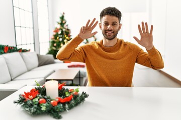 Arab young man sitting on the table by christmas tree showing and pointing up with fingers number nine while smiling confident and happy.