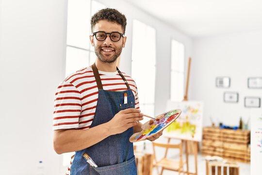 Young arab artist man smiling happy holding paintbrush and palette at art studio.