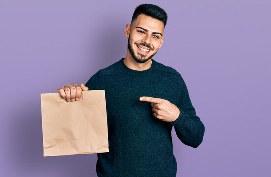 Young hispanic man with beard holding take away paper bag smiling happy pointing with hand and finger