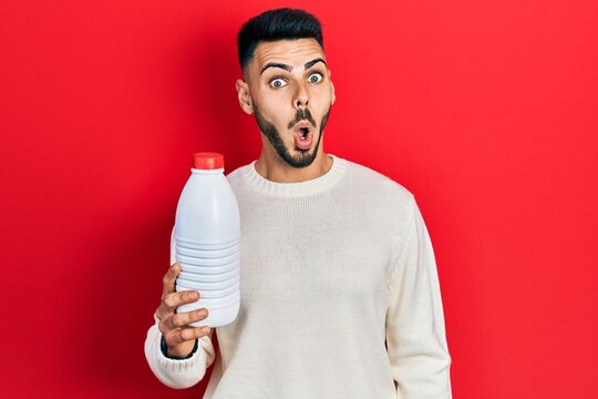 Young hispanic man with beard holding liter bottle of milk scared and amazed with open mouth for surprise, disbelief face
