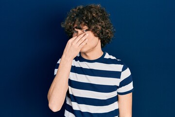 Handsome young man wearing casual striped t shirt smelling something stinky and disgusting, intolerable smell, holding breath with fingers on nose. bad smell