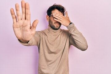 Handsome man with beard wearing turtleneck sweater and glasses covering eyes with hands and doing stop gesture with sad and fear expression. embarrassed and negative concept.