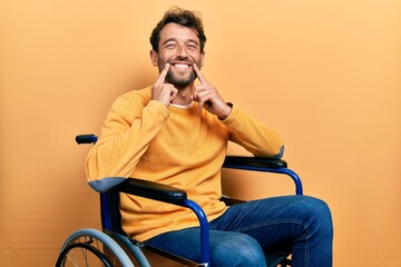 Handsome man with beard sitting on wheelchair smiling with open mouth, fingers pointing and forcing...
