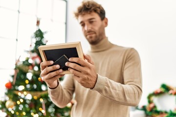 Young hispanic man holding photo standing by christmas tree at home