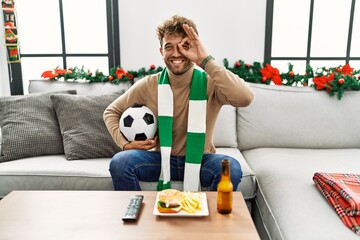 Young hispanic man football hooligan holding ball at home smiling happy doing ok sign with hand on eye looking through fingers