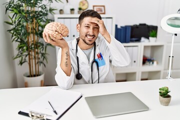 Young doctor holding brain at medical clinic stressed and frustrated with hand on head, surprised and angry face