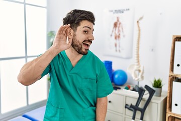 Young man with beard working at pain recovery clinic smiling with hand over ear listening an hearing to rumor or gossip. deafness concept.