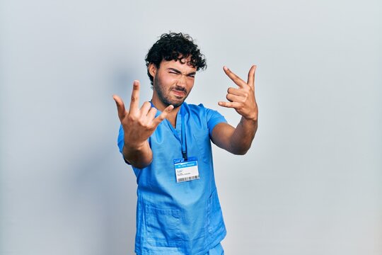 Young hispanic man wearing blue male nurse uniform shouting with crazy expression doing rock symbol with hands up. music star. heavy concept.
