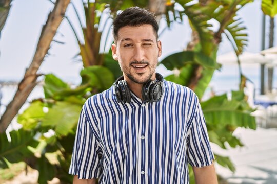 Young handsome man listening to music using headphones outdoors winking looking at the camera with sexy expression, cheerful and happy face.